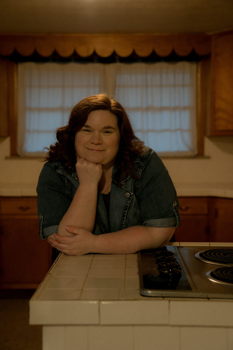 a woman in a green dress and jean jacket stands in an empty kitchen